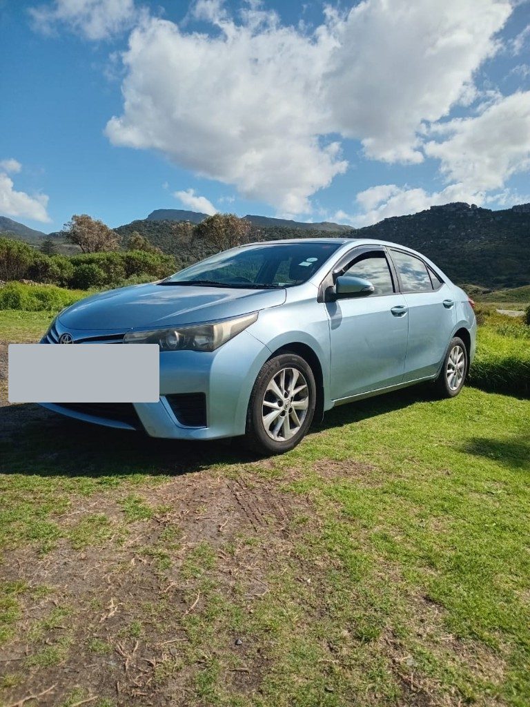 Light blue Toyota Corolla parked on grass with mountains behind it