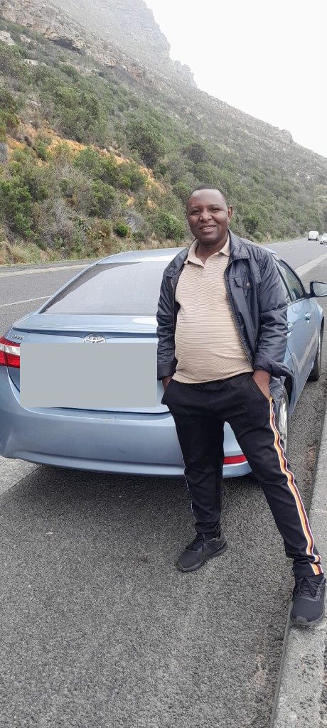 Fanuel Chinovunye standing with his light blue Toyota Corolla on a mountain road